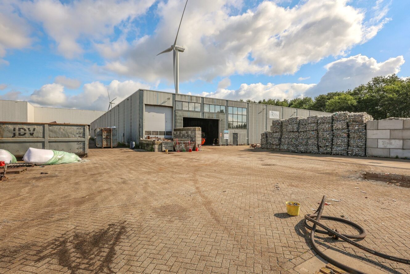 Industrieterrein met groot metalen magazijn, gestapelde balen recyclebare materialen, betonblokken en een zichtbare windturbine onder een gedeeltelijk bewolkte hemel.