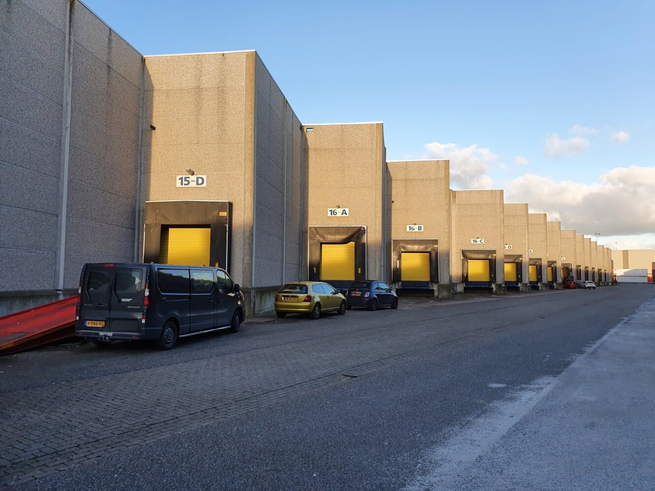 Een rij laadkades van industriële magazijnen met gesloten gele deuren en verschillende geparkeerde voertuigen op een verharde oprit.