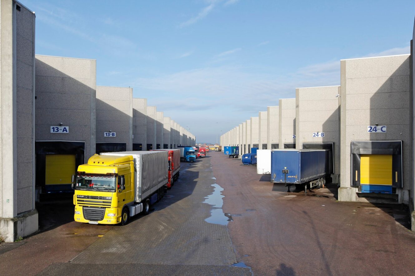Rijen laadkades van magazijnen met vrachtwagens, waaronder een prominente gele vrachtwagen, geparkeerd bij verschillende kades op een bewolkte dag.