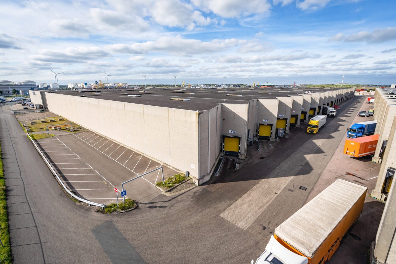Een groot magazijn met meerdere laadkades, verschillende vrachtwagens geparkeerd om te laden en lossen en een aangrenzend leeg parkeerterrein onder een gedeeltelijk bewolkte hemel.