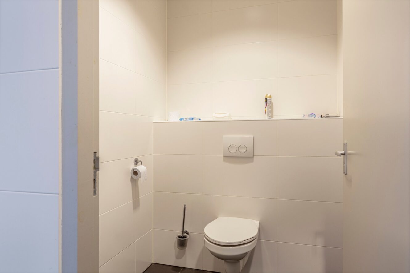 Een kleine badkamer met een aan de muur bevestigd toilet, toiletpapierhouder, toiletborstel en persoonlijke verzorgingsproducten op een plank tegen wit betegelde muren.