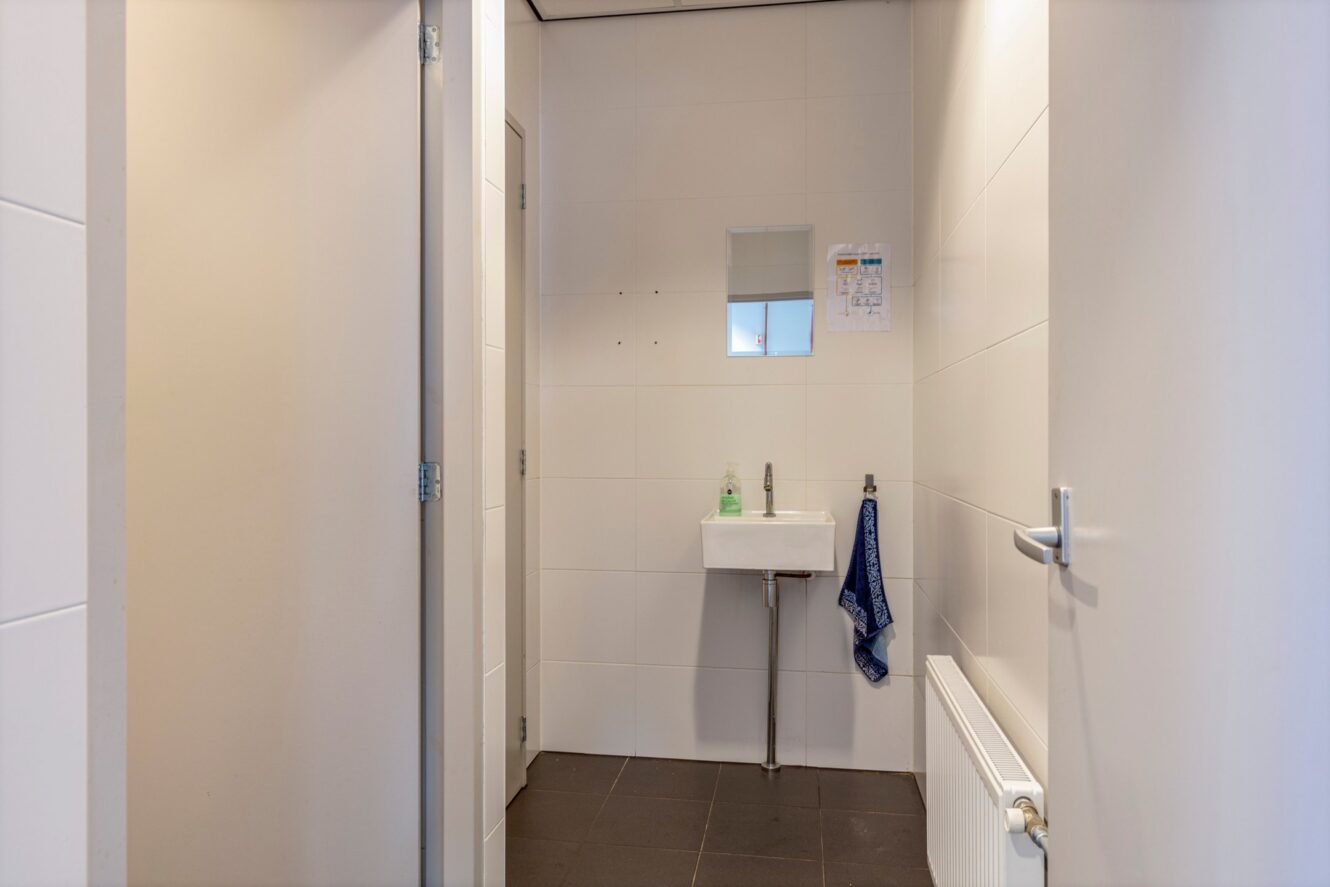 Een klein, eenvoudig toilet met wit betegelde muren, een aan de muur bevestigde wastafel, een spiegel, een handdoek, zeep en een radiator onder een aan de muur bevestigde kalender.