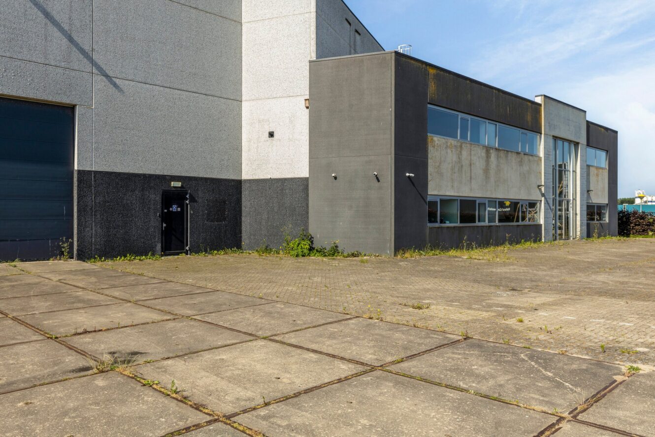 Een groot, grijs industrieel gebouw met meerdere ramen, een gesloten garagedeur, betonnen stoeptegels en wat onkruid dat tussen de kieren groeit.