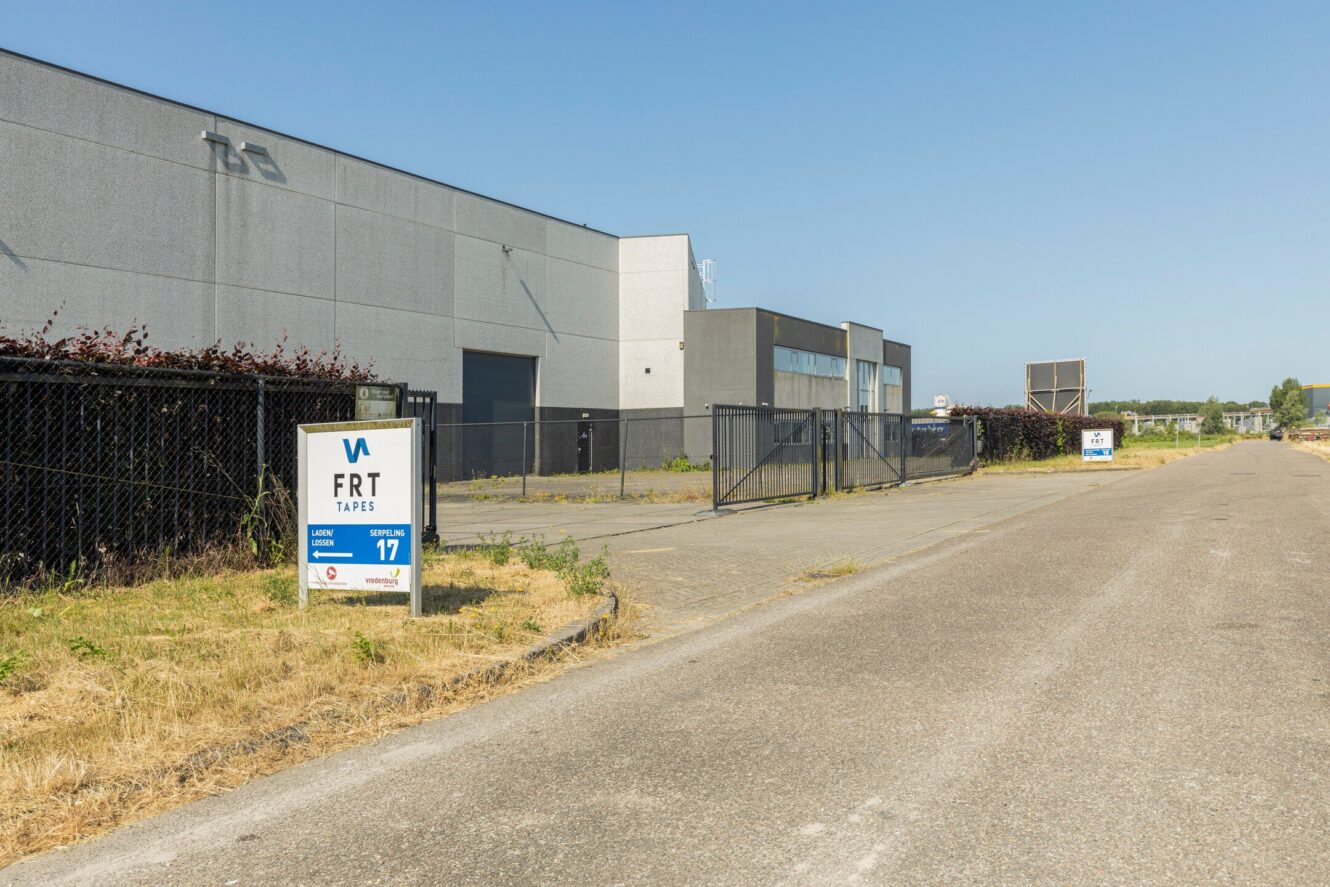 Industrieel gebouw met grijze gevel en omheind terrein; borden bij de ingang geven FRT Tapes aan met adresnummer 17 op een zonnige dag.