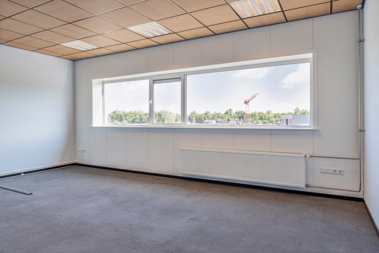 Lege kantoorkamer met een groot raam, beige plafondpanelen, witte muren, muurradiator en grijze tapijtvloer. Buiten is bouwmateriaal zichtbaar.