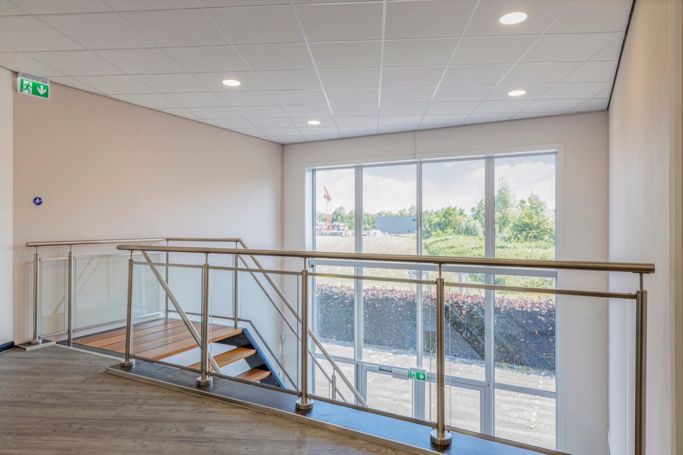 Moderne binnentrap met metalen leuningen, grote ramen die natuurlijk licht binnenlaten en zichtbare groene bordjes met nooduitgangen op het plafond en de deur.