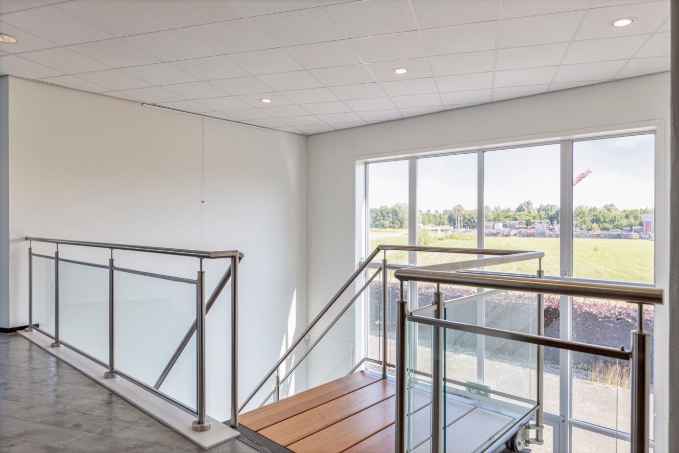 Binnentrap met metalen leuningen en houten treden, naast grote ramen met uitzicht op een grasveld buiten met bomen en een bouwkraan in de verte.