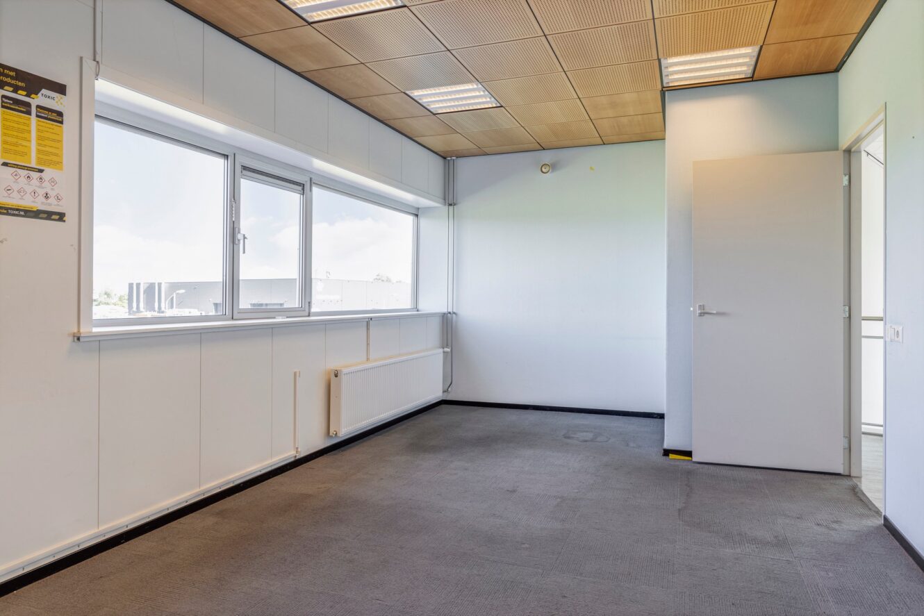 Een lege, ongemeubileerde kantoorkamer met grote ramen, een radiator, een open deur en een licht houten panelenplafond. Op de linkermuur is een veiligheidsposter te zien.