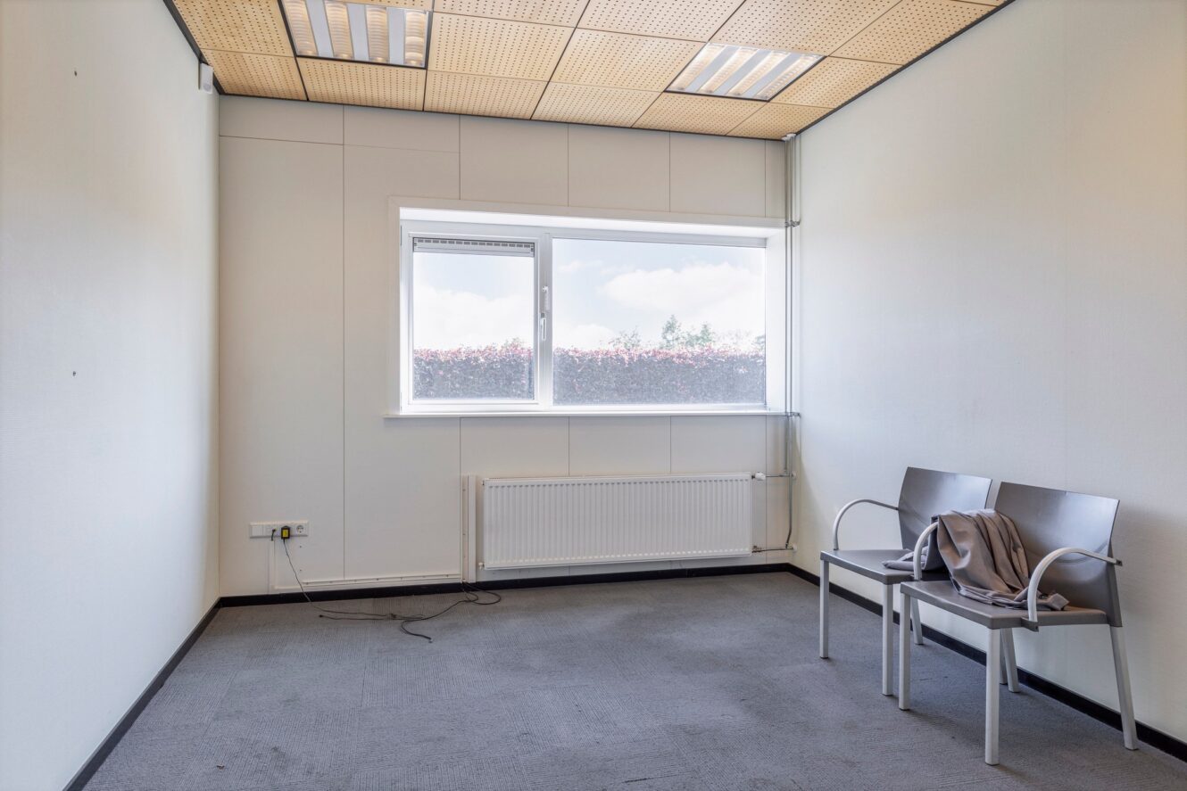 Een grotendeels lege kantoorkamer met gladde muren, een raam, TL-verlichting aan het plafond, een radiator en twee metalen stoelen met een jas over één ervan gedrapeerd. Een netsnoer ligt op de vloer.