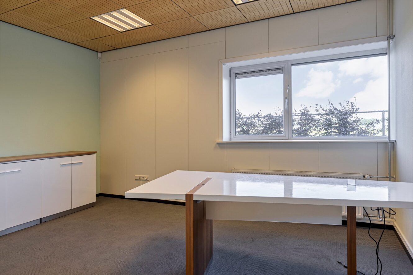 Een lege kantoorkamer met een wit bureau, witte opbergkasten, beige muren, een groot raam en grijze vloerbedekking.