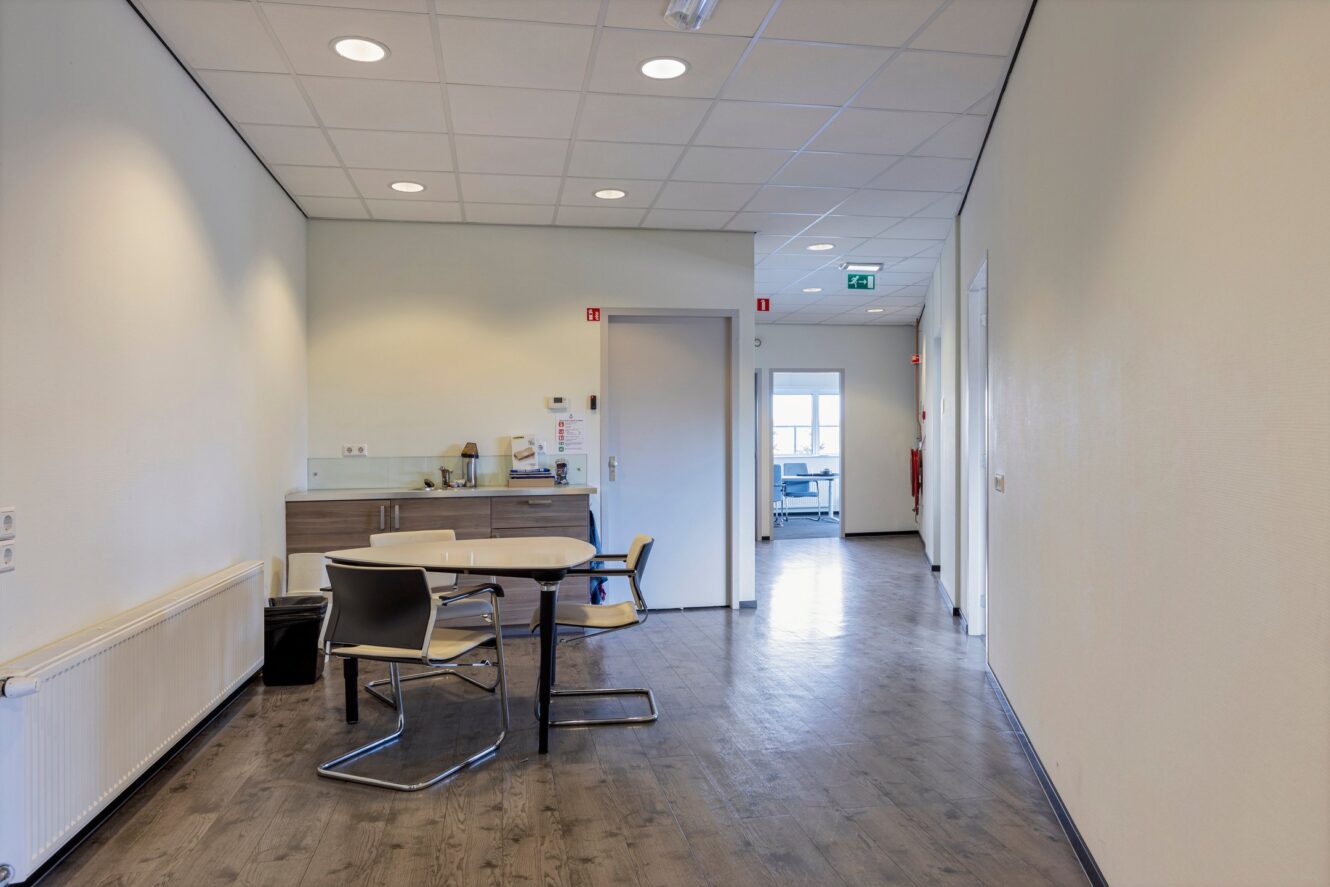 Een gang met een kleine tafel en stoelen, een keukentje aan de linkerkant en een open deur die leidt naar een andere kantoorruimte op de achtergrond.