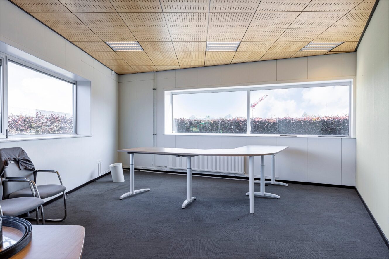 Leeg kantoor met een groot L-vormig bureau, drie stoelen, een prullenbak en grote ramen die daglicht binnenlaten.