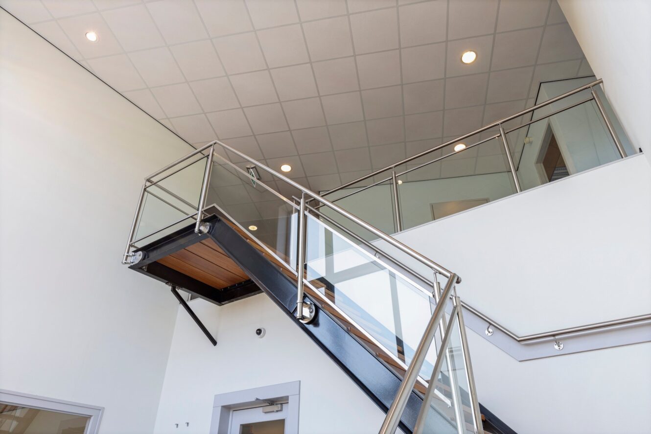 Moderne binnentrap met glazen en metalen leuningen die leidt naar een bovenverdieping in een gebouw met witte muren en een plafond van druppeltegels met inbouwverlichting.