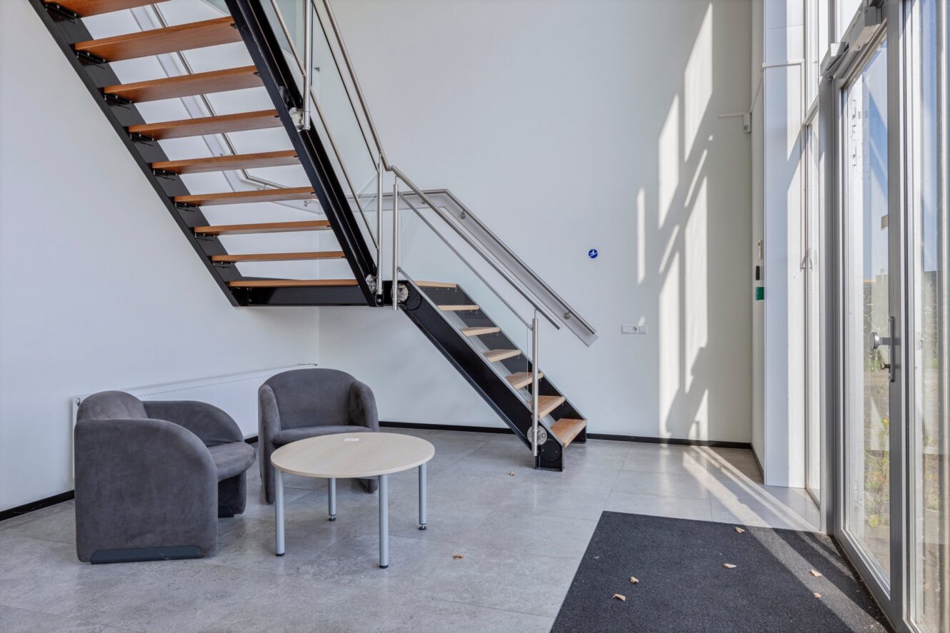 Een modern interieur met een metalen en houten trap, twee grijze stoelen en een ronde tafel op een tegelvloer bij grote ramen.