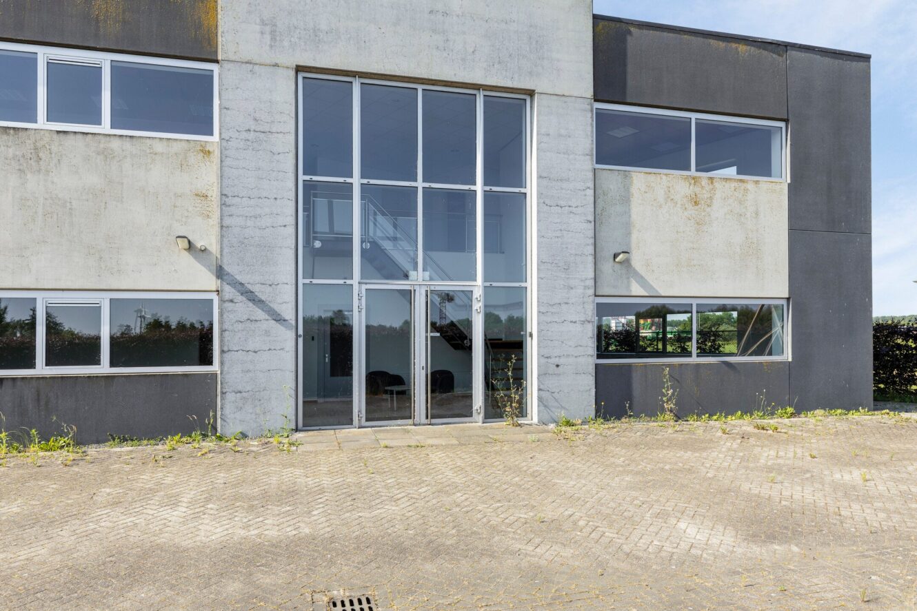 Vooraanzicht van een modern commercieel gebouw van twee verdiepingen met grote glazen toegangsdeuren, betonnen gevel, kleine ramen en een geplaveid gedeelte aan de voorkant.