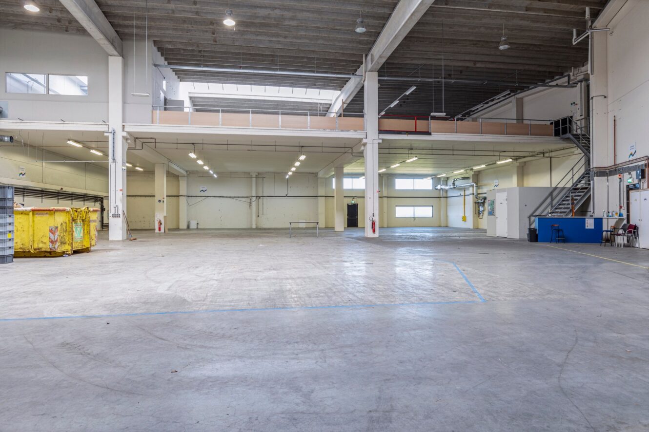Ruim leeg magazijn met betonnen vloeren, hoge plafonds, bovenverlichting, gele bakken, blauwe container en een metalen trap naar een tussenverdieping.