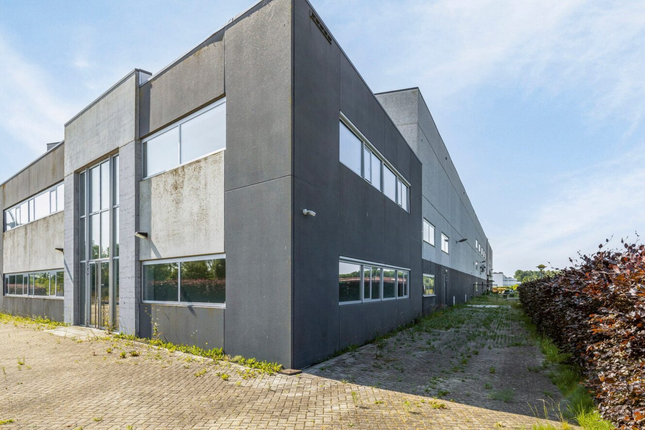 Een modern, grijs industrieel gebouw met grote ramen, omgeven door geplaveide en grasvelden onder een gedeeltelijk bewolkte hemel.