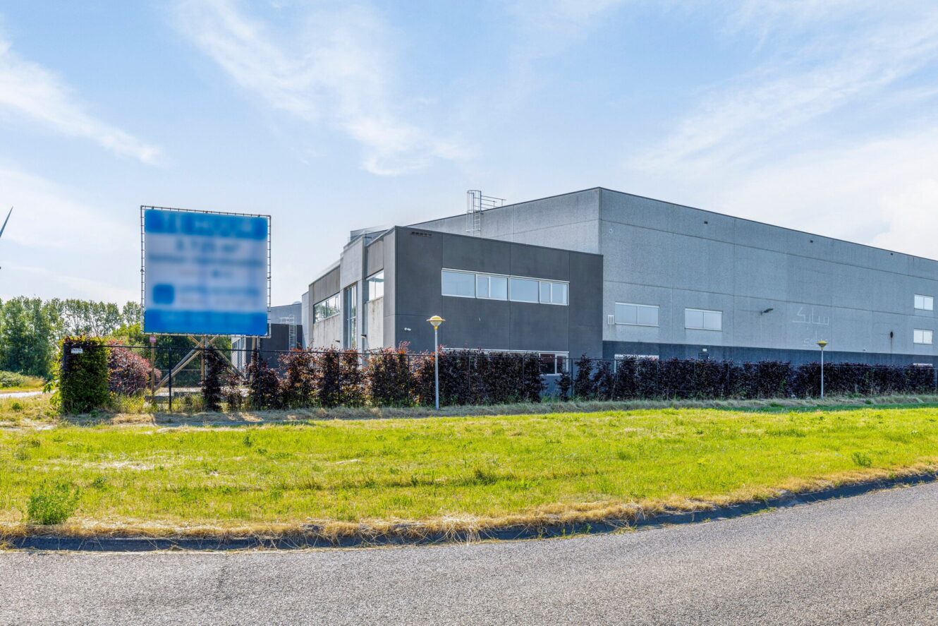 Een groot, modern industrieel gebouw met een leeg blauw bord ervoor, omringd door gras, struiken en een bochtige weg onder een gedeeltelijk bewolkte hemel.