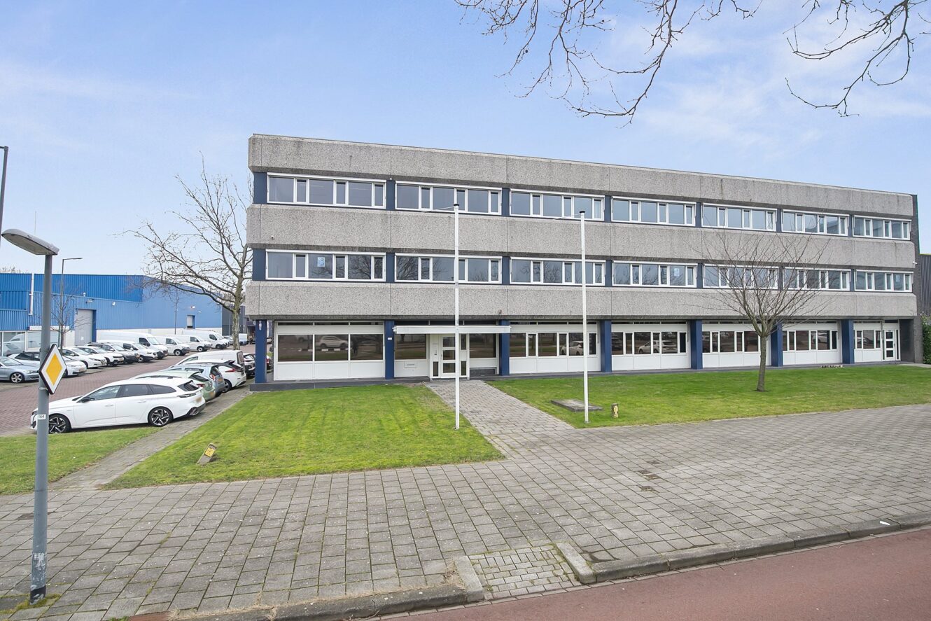 Betonnen kantoorgebouw van drie verdiepingen met grote ramen, een geplaveid trottoir en geparkeerde auto's aan de linkerkant; bomen zonder bladeren en gras aan de voorkant.