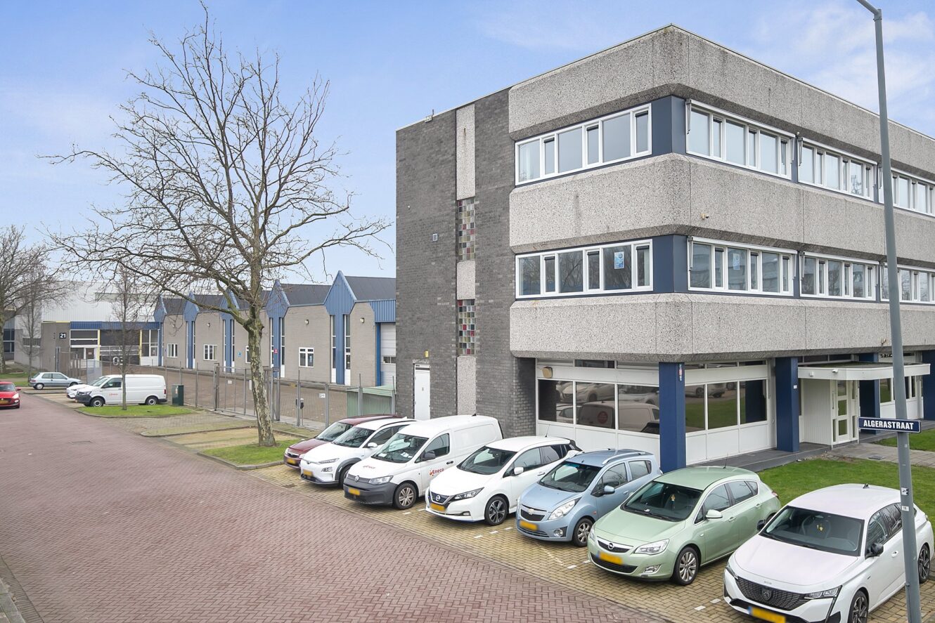 Een kantoorgebouw van drie verdiepingen met geparkeerde auto's ervoor, gelegen in een rustige straat met soortgelijke gebouwen en een boom zonder bladeren in de buurt.