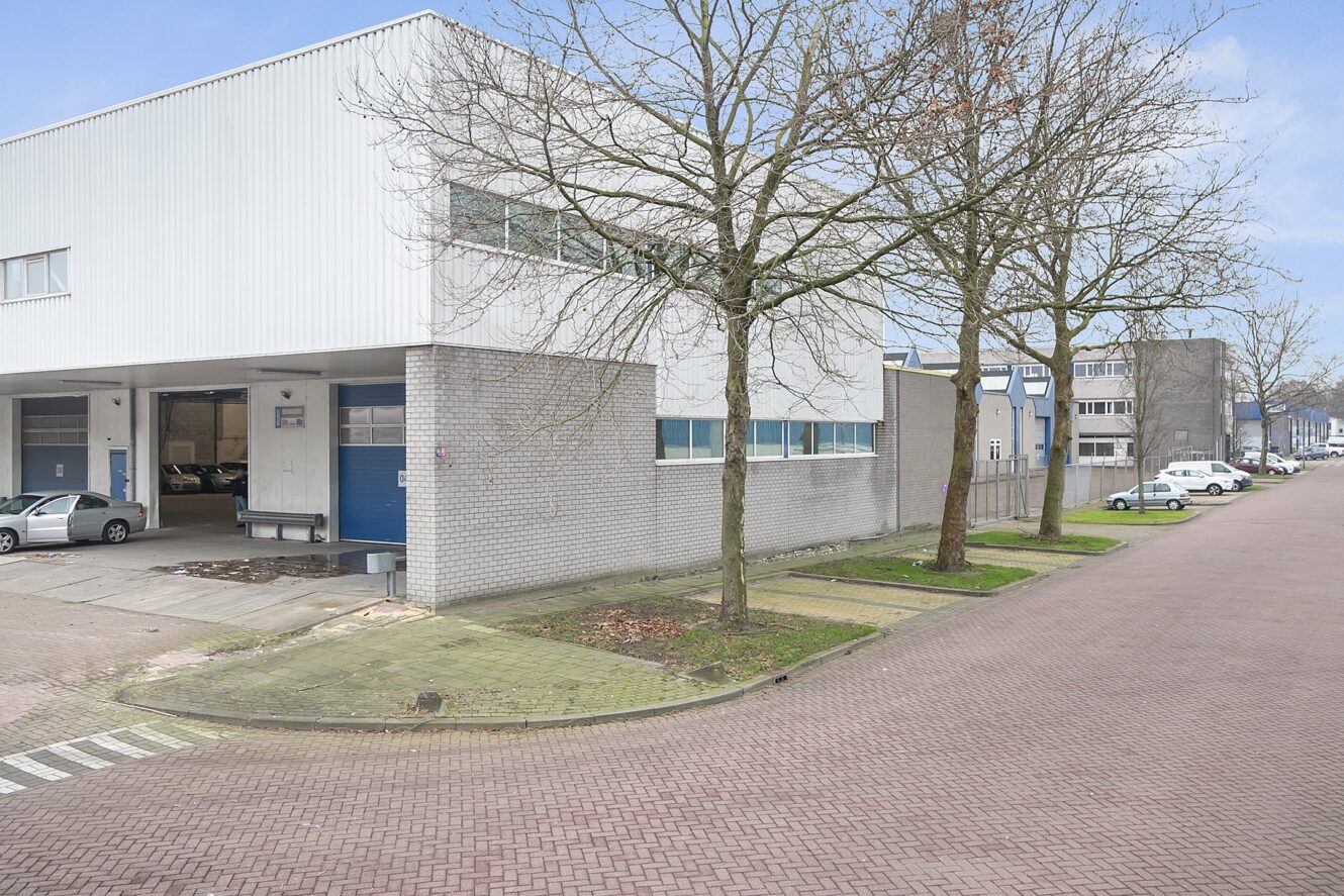 Een modern industrieel gebouw met een grijze bakstenen buitenkant en blauwe accenten ligt aan een rustige straat met bladerloze bomen en geparkeerde auto's.