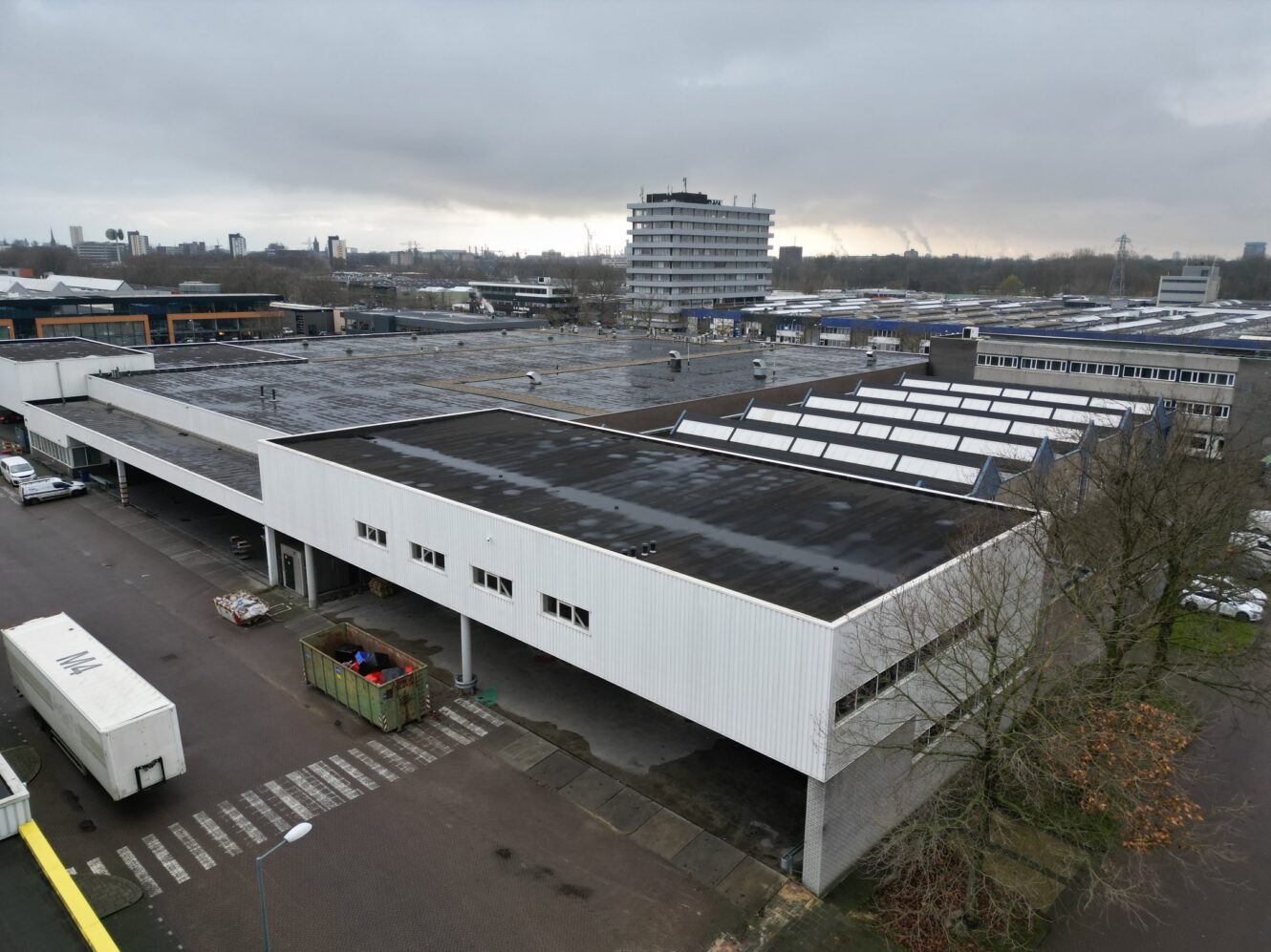 Luchtfoto van een industrieel pakhuiscomplex met platte daken, omringd door andere gebouwen, geparkeerde vrachtwagens en een bewolkte lucht.
