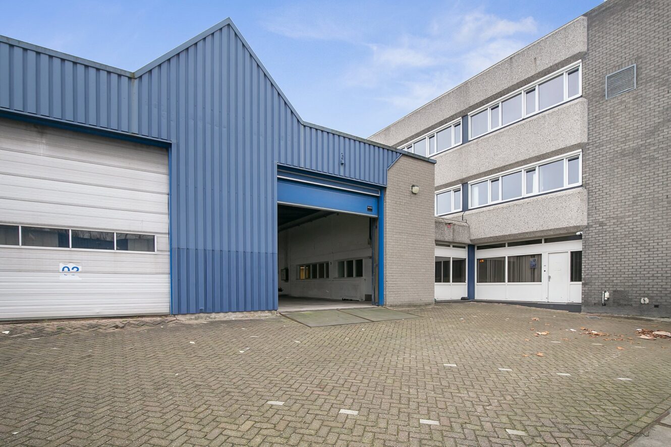 Industrieel gebouw met een blauwe metalen gevel, grote garagedeuren en aangrenzende grijze bakstenen structuur, op een geplaveid perceel onder een bewolkte hemel.