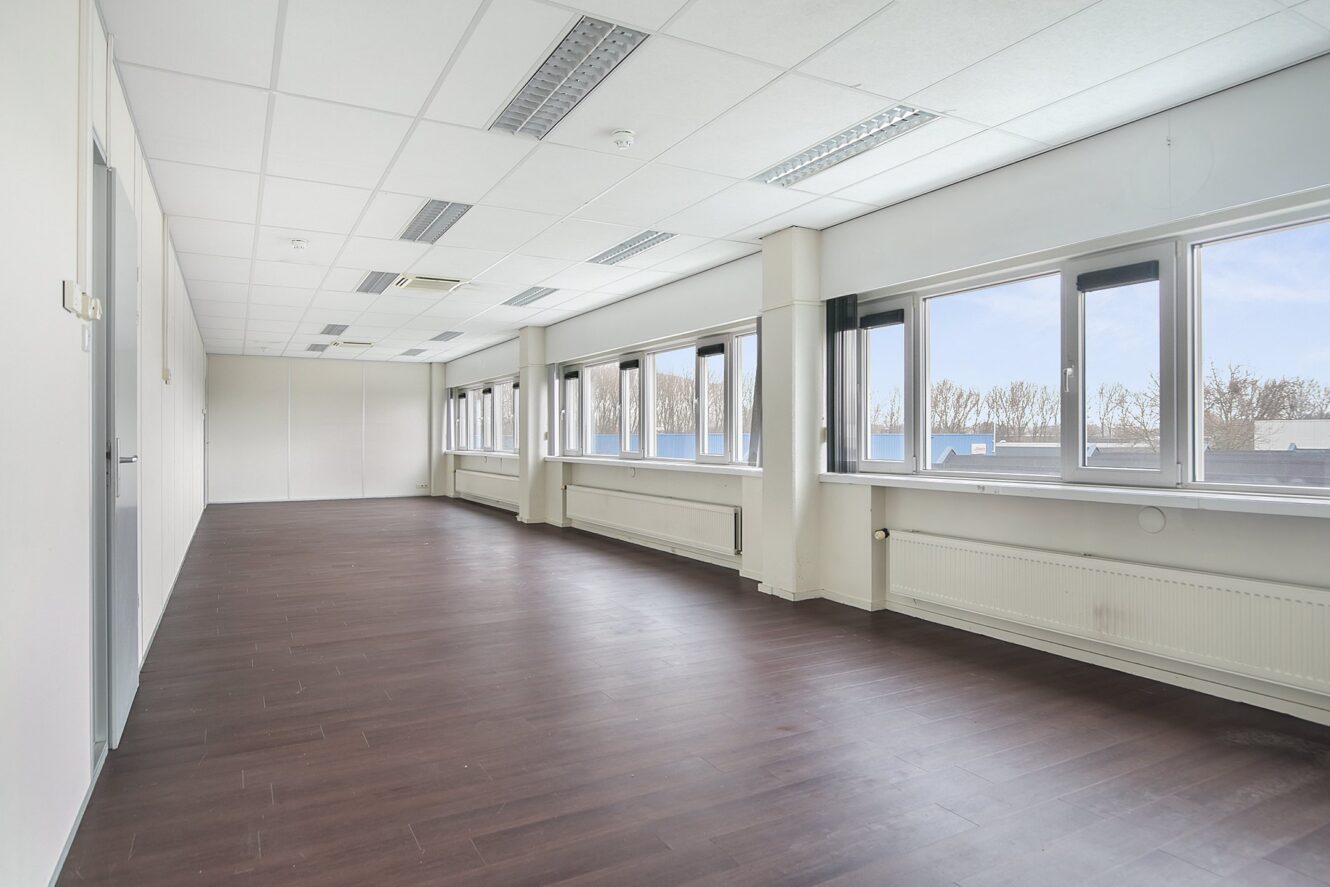Een ruime, lege kantoorkamer met een donkerhouten vloer, witte muren, een verlaagd plafond met verlichting en grote ramen die natuurlijk licht binnenlaten.