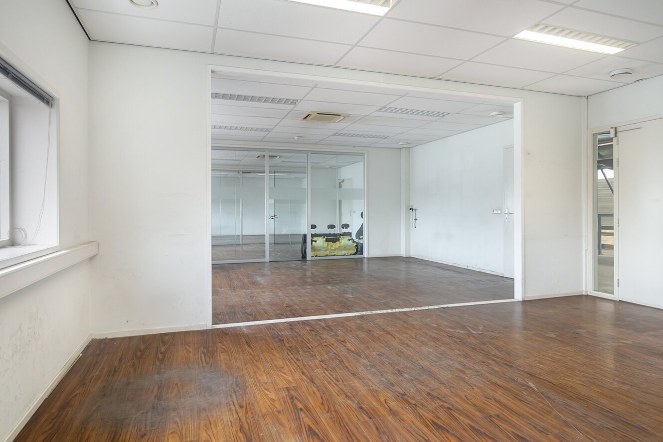 Lege kantoorruimte met witte muren, houten vloeren, een glazen wand en plafondverlichting. Aan de linkermuur zit een klein raam en rechts zitten twee gesloten deuren.