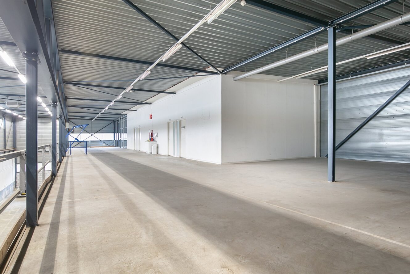 Leeg interieur van een industrieel pakhuis met betonnen vloer, metalen balken en een witte ruimte met scheidingswanden; zonlicht stroomt door de ramen, brandblussers aan de muur.