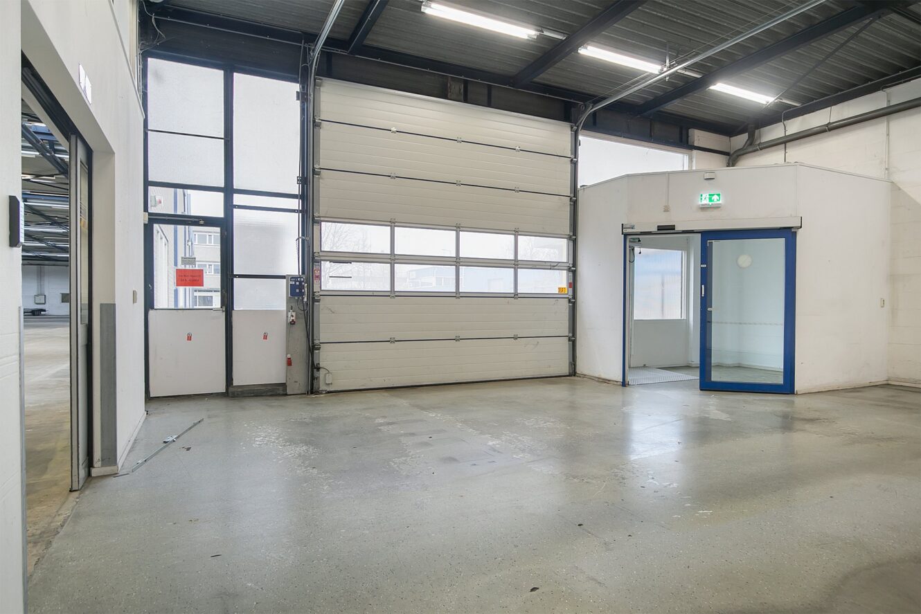 Interieur van een leeg industrieel pakhuis met een grote garagedeur, glazen toegangsdeur met blauw frame en gepolijste betonnen vloer.