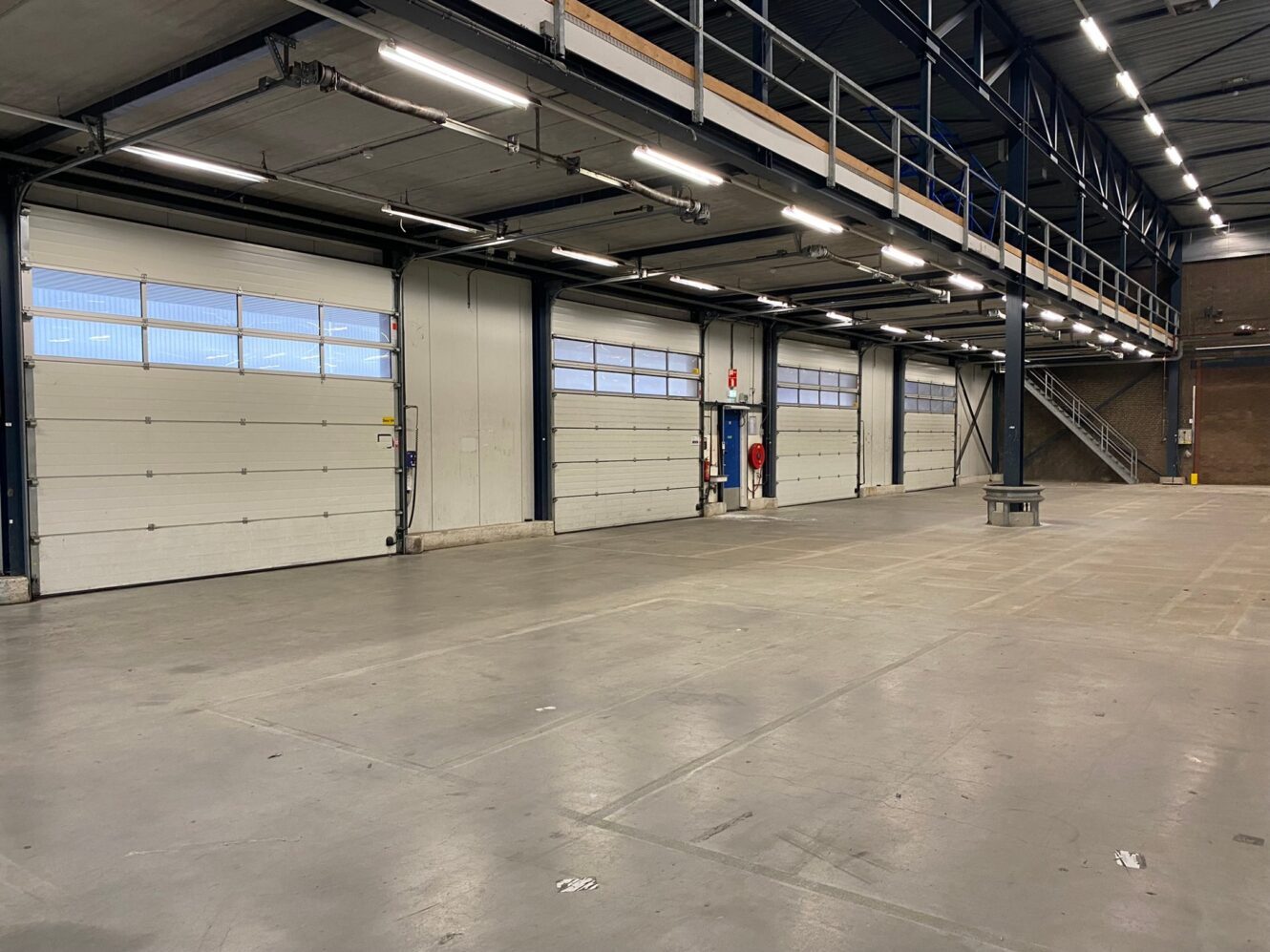 Groot, leeg industrieel pakhuis met vier witte oprolbare garagedeuren, betonnen vloer, bovenlichten, een trap en een tussenverdieping.