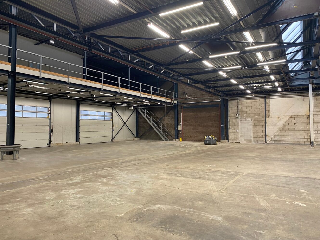 Groot, leeg magazijninterieur met betonnen vloer, metalen balken, bovenverlichting, een trap naar een mezzanine en grote garagedeuren langs één muur.