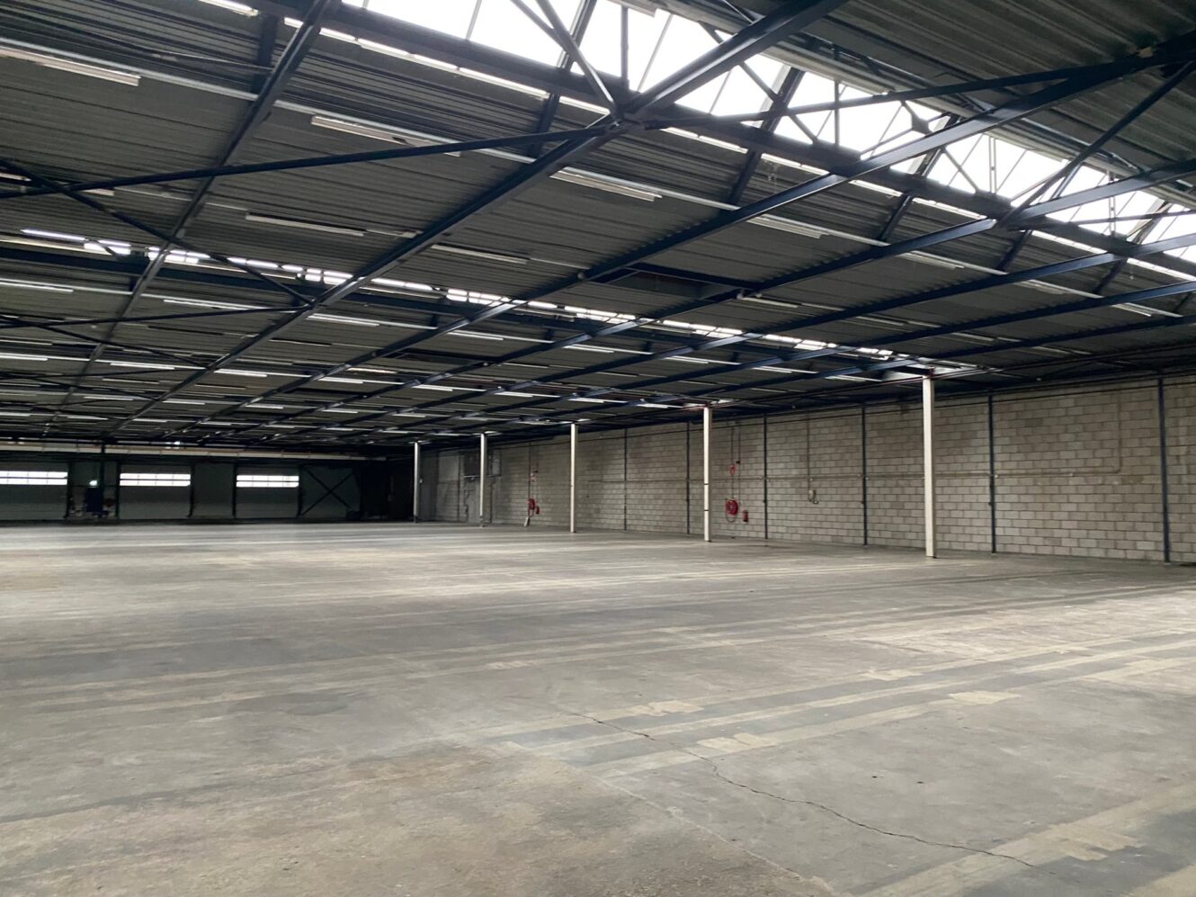 Groot, leeg industrieel pakhuis met betonnen vloeren, zichtbare metalen balken en bakstenen muren, verlicht door natuurlijk licht van dakramen.