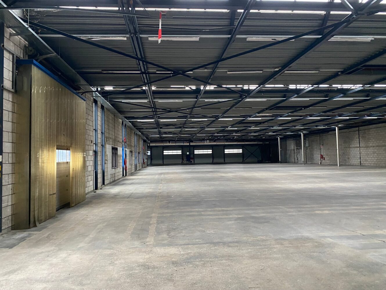 Groot, leeg pakhuis met betonnen vloeren, hoog plafond, zichtbare balken, muren van sintelblokken en wat licht door ramen en deuren aan de linkerkant.