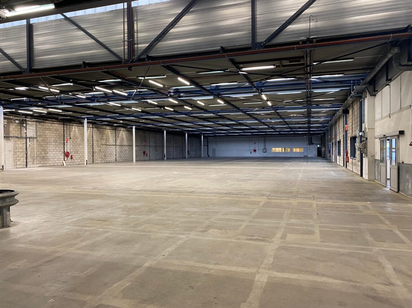 Groot, leeg industrieel pakhuis met betonnen vloer, zichtbare plafondbalken en bakstenen muren. Fluorescentielampen aan het plafond. Er zijn geen apparaten of mensen aanwezig.