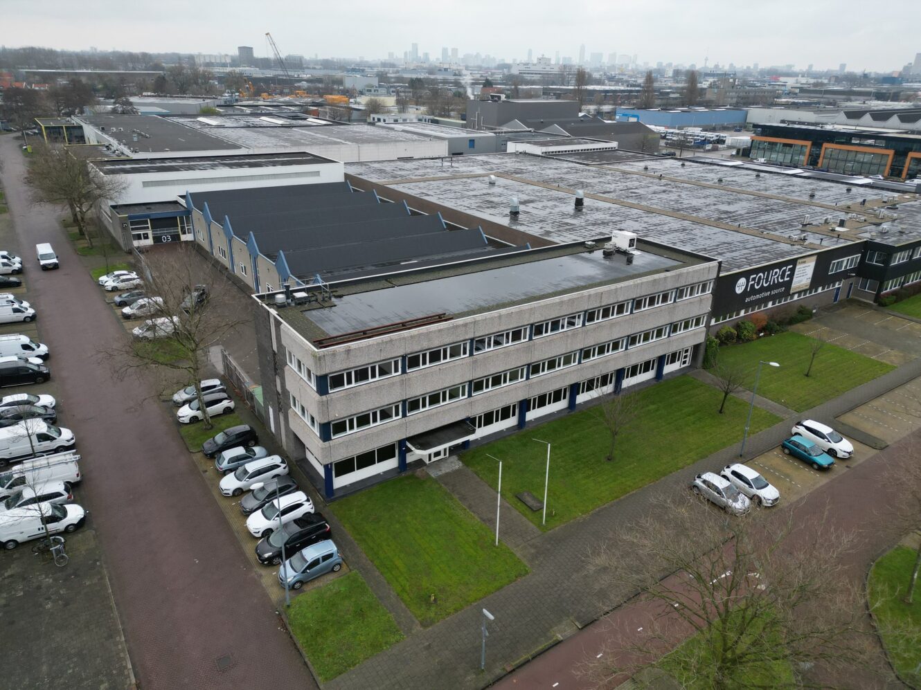 Luchtfoto van een industrieel kantoorgebouw met plat dak, aangrenzende parkeerplaatsen en omliggende commerciële gebouwen op een bewolkte dag.