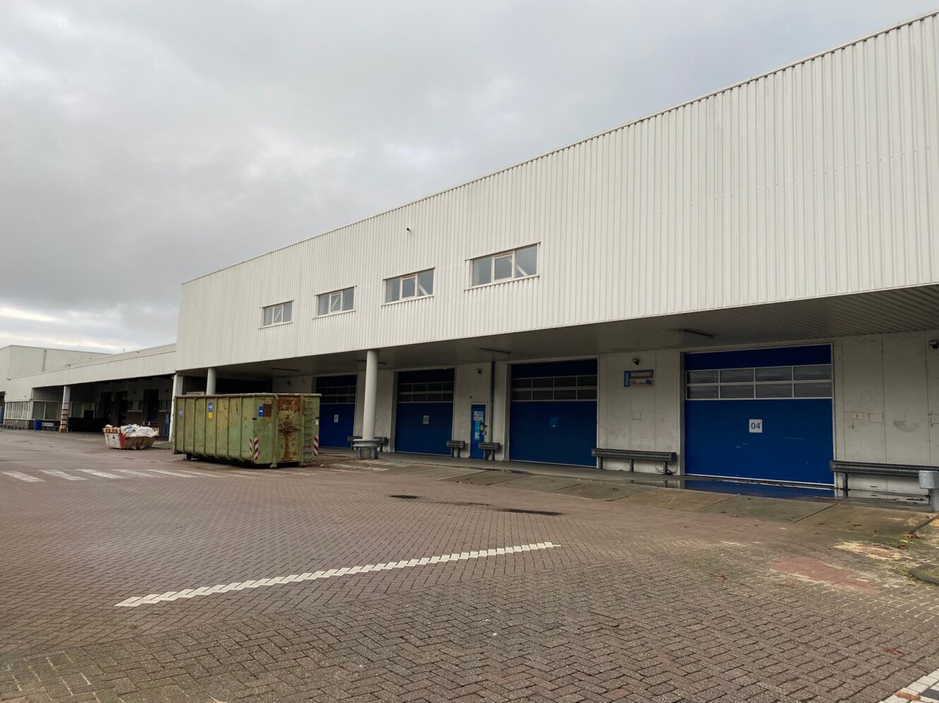 Een groot industrieel gebouw met verschillende blauwe laadperrondeuren, een gele afvalcontainer en een leeg geplaveid terrein ervoor onder een bewolkte hemel.