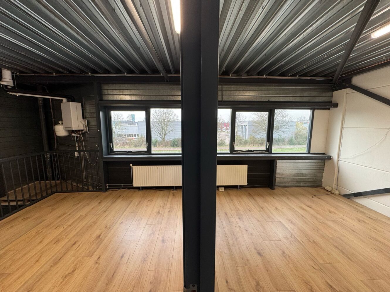 Een gedeelde ruimte in industriële stijl met houten vloeren, grote ramen, radiatoren en een centrale metalen steunbalk, met uitzicht op een landschap buiten.