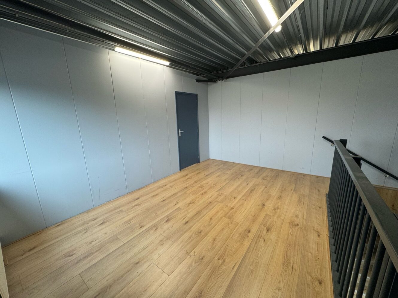 Lege kamer met lichthouten vloer, muren met grijze panelen, een gesloten grijze deur, metalen plafond en zwarte leuning naast een trap.