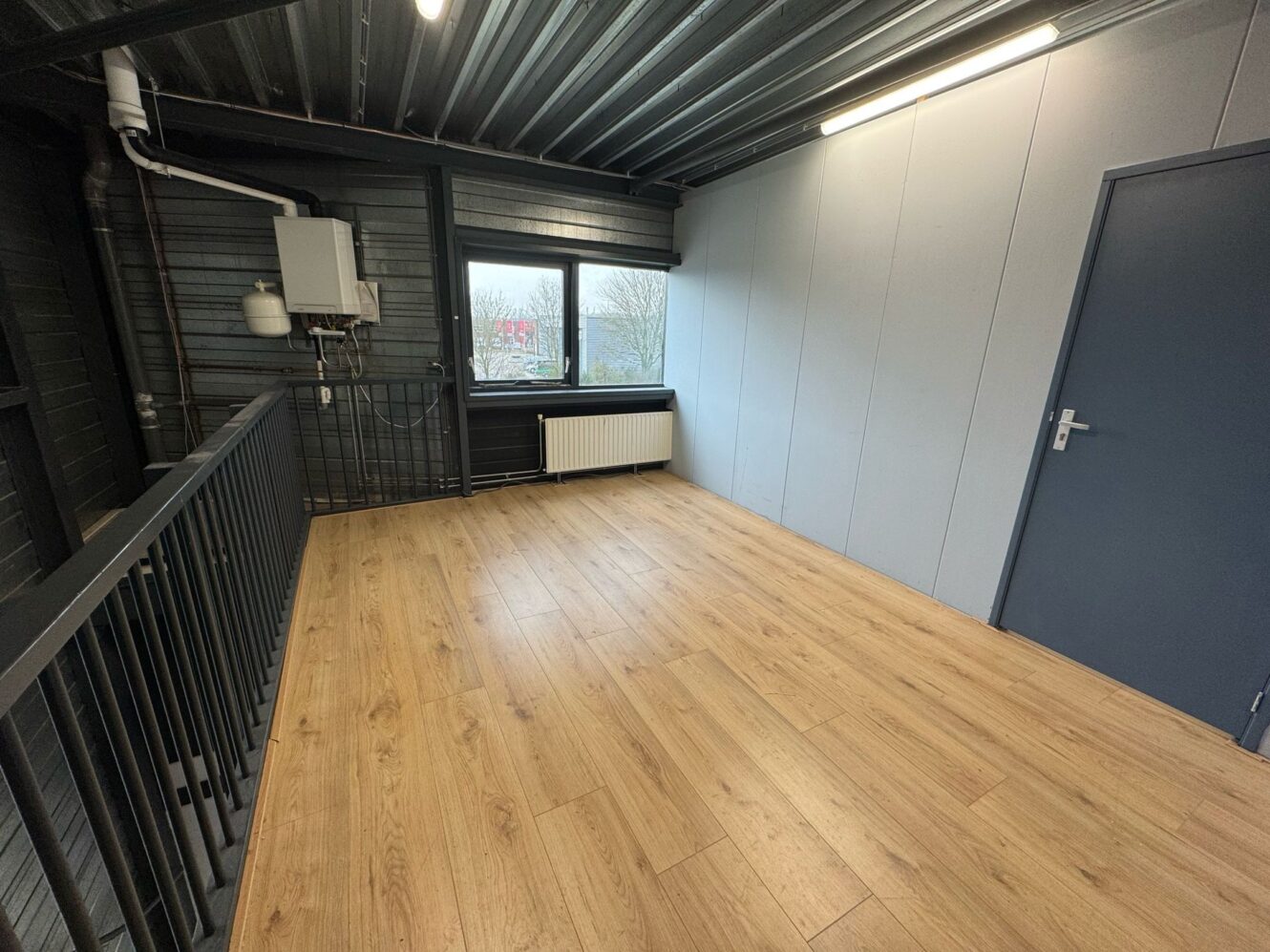 Kleine, lege kamer met een licht houten vloer, een grijs metalen plafond, een radiator onder een raam, zichtbare leidingen aan de muur en een gesloten blauwe deur.