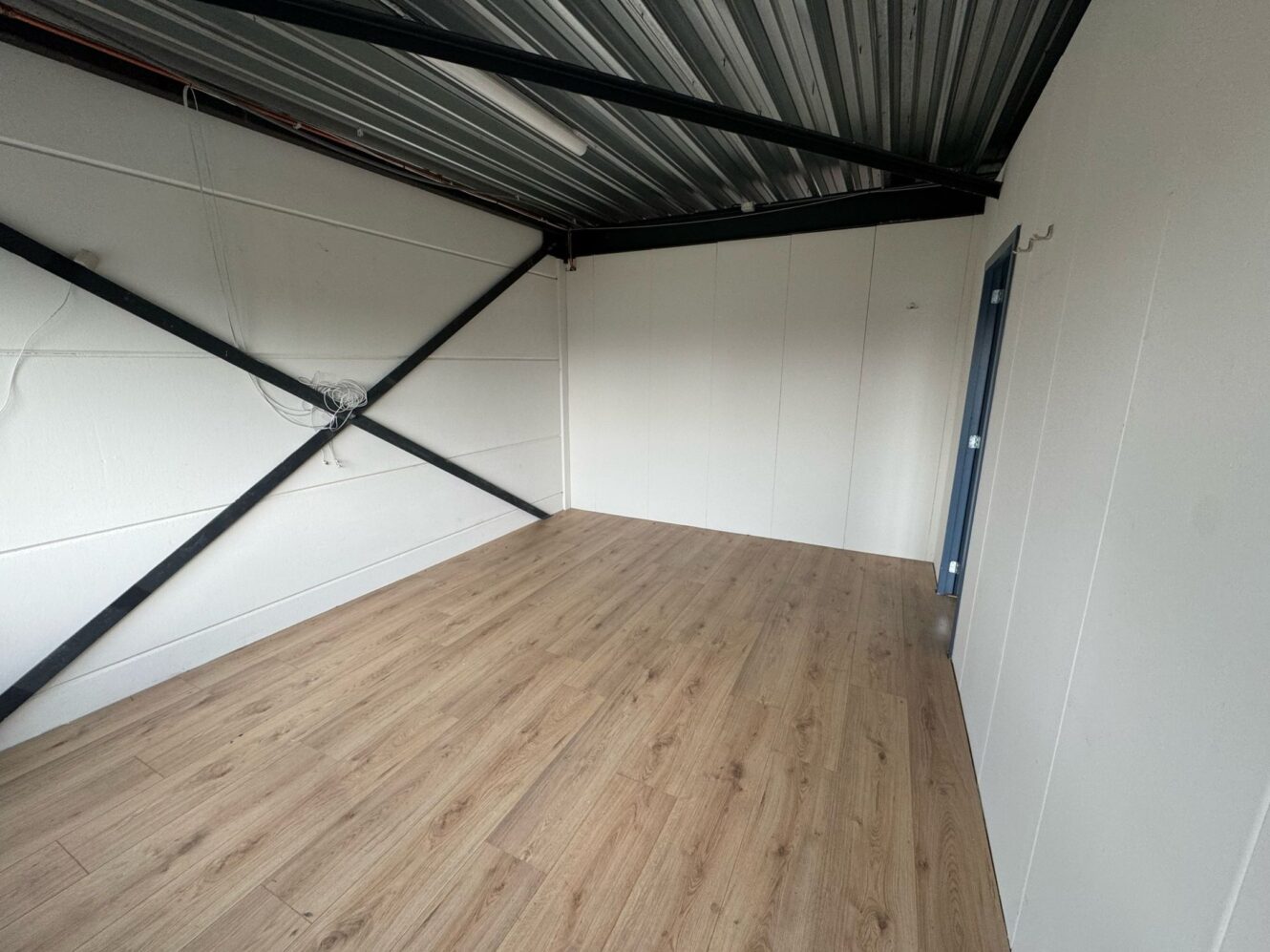 Lege kamer met witte muren, een metalen plafond met zichtbare balken, houten vloeren en een deur met blauw frame aan de rechterkant.