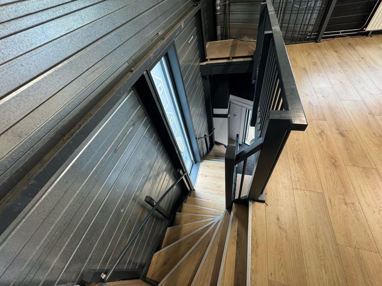 Houten wenteltrap met zwarte metalen leuning die naar beneden leidt vanuit een zolderruimte met lichte houten vloeren en donkere wanden met panelen.