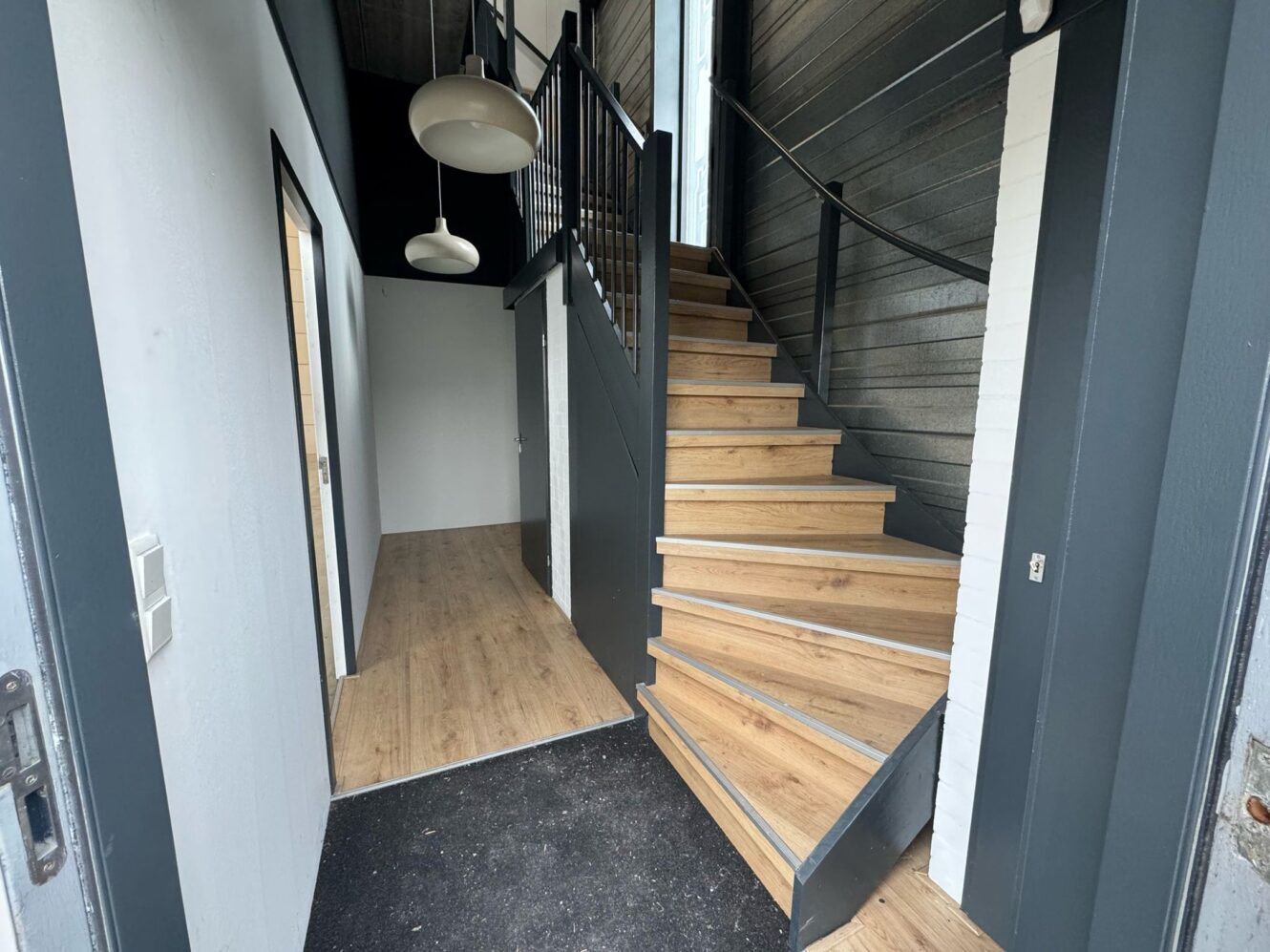 Moderne hal met houten trap naar boven, lichte houten vloer, witte en donkergrijze muren en twee ronde plafondlampen. Aan de linkerkant zijn twee open deuropeningen zichtbaar.