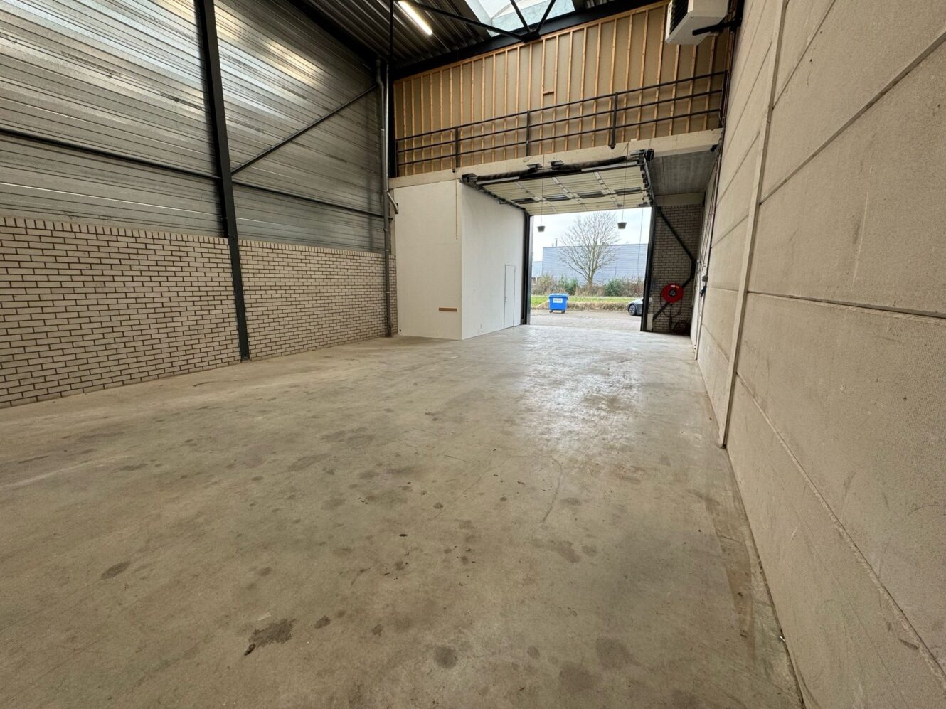Leeg industrieel magazijn met betonnen vloer, bakstenen en paneelmuren, een hoog plafond en een open roldeur die naar buiten leidt.