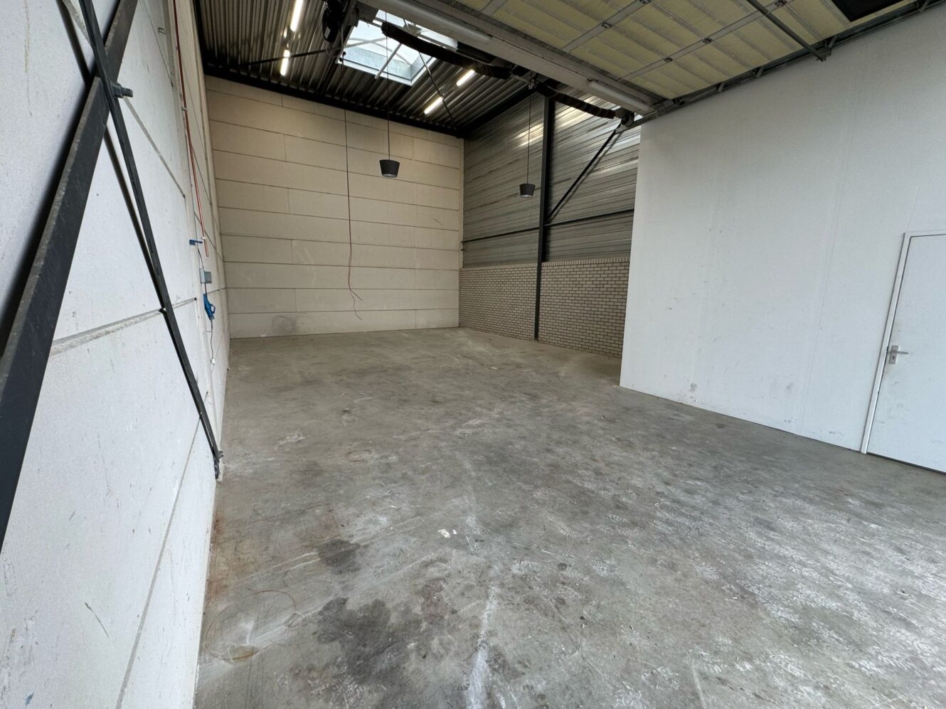 Leeg industrieel pakhuis met betonnen vloer, hoog plafond, dakraam, metalen balken en gedeeltelijk open garagedeur aan de rechterkant.
