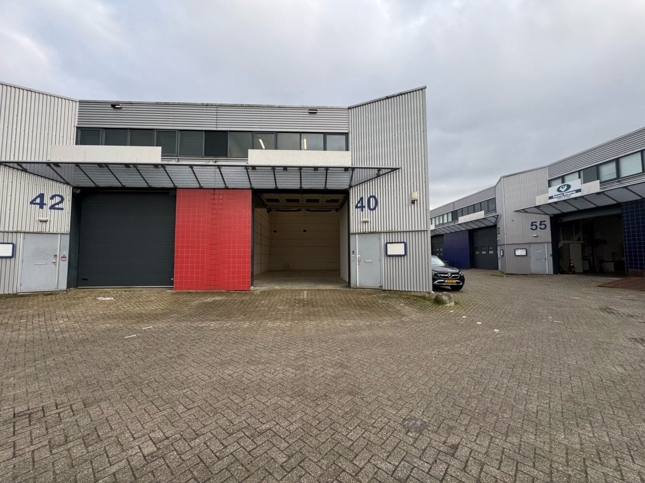 Industriële magazijnunits met nummers 40, 42 en 55 zichtbaar; unit 40 heeft een open garagedeur en een geparkeerde zwarte auto ernaast. De lucht is bewolkt.