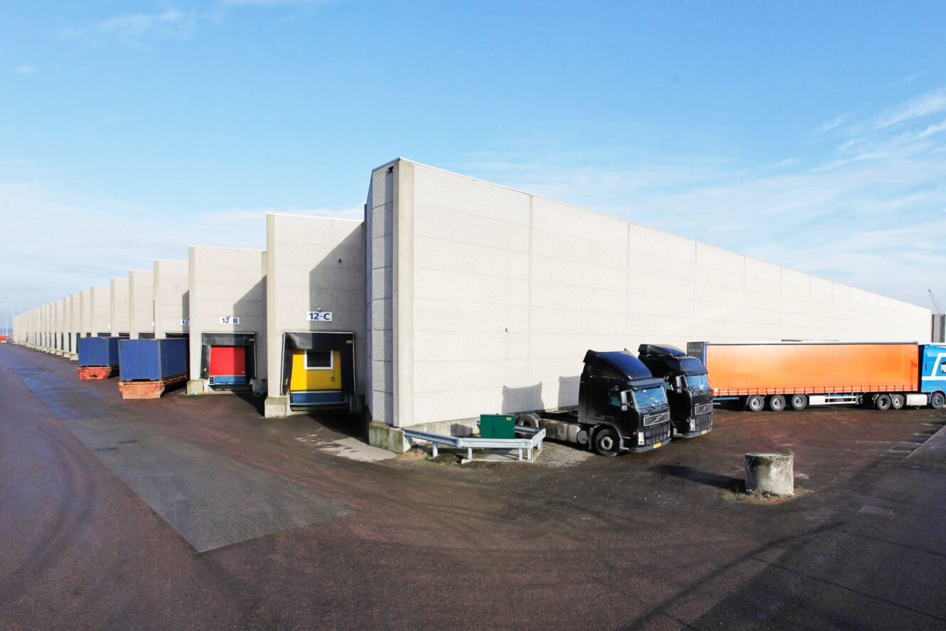 Een rij laadkades van een magazijn met geparkeerde vrachtwagens en opleggers op een heldere dag.