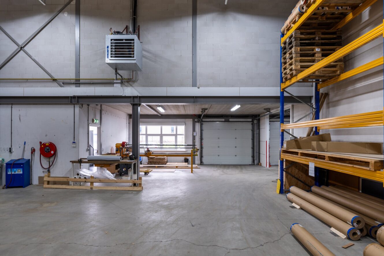 Interieur van een ruim magazijn met betonnen vloeren, rekken, werkbanken, gestapelde pallets en grote rollen papier. Een garagedeur en industriële apparatuur zijn zichtbaar.