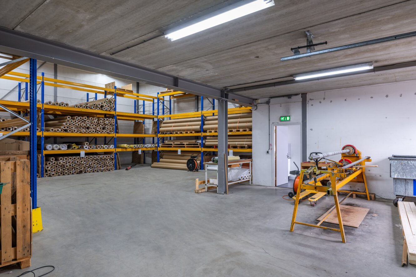 Magazijninterieur met industriële rekken voor grote rollen materiaal, verspreide apparatuur en TL-verlichting boven het hoofd.